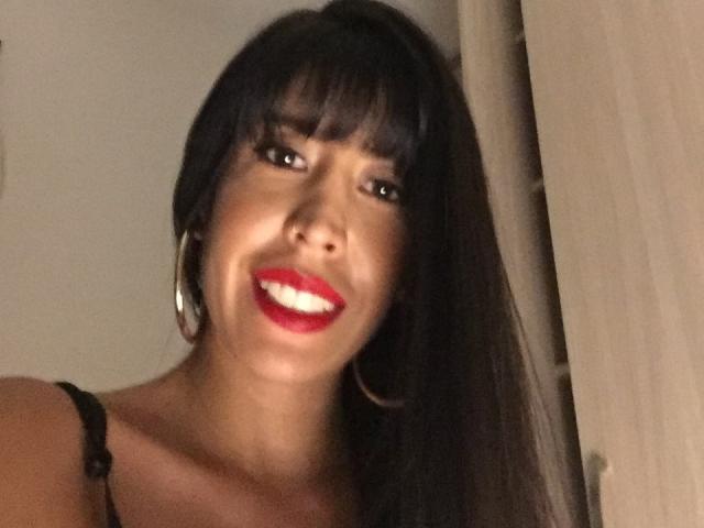 SeleneBrigth with dark, straight hair, bangs, and red lipstick, smiling, wearing black straps and hoop earrings, showing her white teeth. Her brown eyes are slightly up.