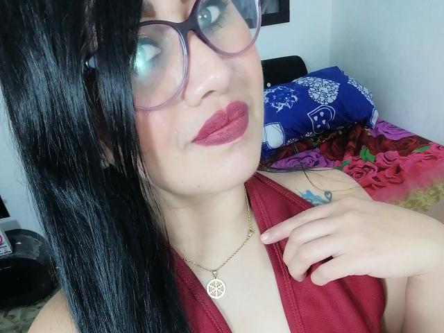 CathyCat mit schwarzen Haaren, Brille, rotem Lippenstift, rotem Kleid, goldener Halskette, die ihre Brüste zeigt, Hand auf der Brust, kleines Tattoo, lächelnd, helle Haut, Schlafzimmer, blaues Kissen im Hintergrund..