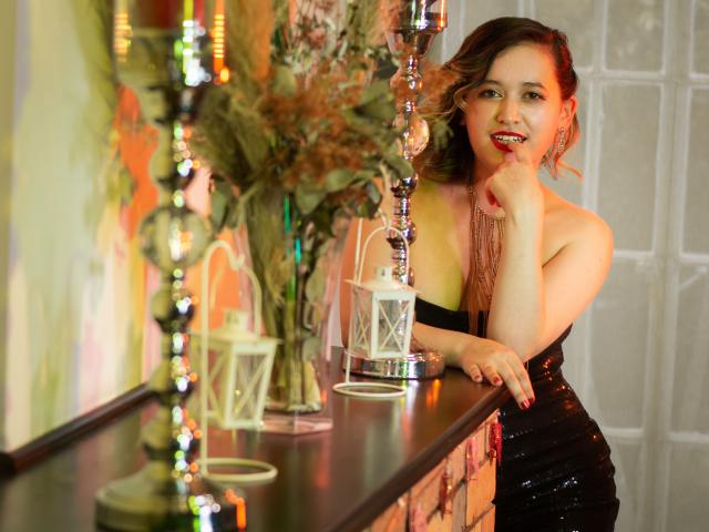 Bianca Cabassi, con cabello castaño ondulado, lápiz labial rojo y un vestido negro con lentejuelas, tocándose la barbilla, junto a un candelabro y faroles blancos sobre una superficie de madera oscura..