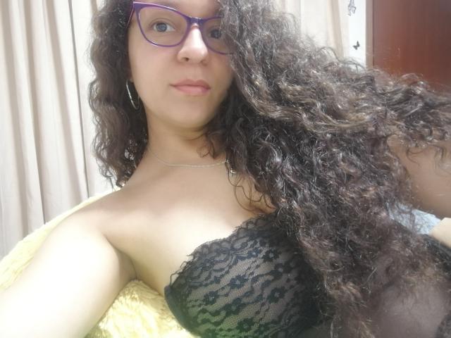 Angelonix with curly black hair, purple glasses, and a black lace strapless top, showing her boobs, yellow blanket, and a silver necklace in a slightly blurry selfie.
