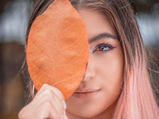 ValerieMartens sosteniendo una gran hoja naranja sobre su ojo derecho, cabello rosa, maquillaje de ojos morado, ligera sonrisa, concentrada en su ojo izquierdo, luz natural suave, expresión gentil..
