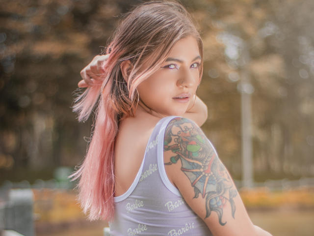 ValerieMartens, com cabelo com pontas rosa, olhando por cima do ombro, mostrando uma tatuagem colorida no braço direito, vestindo uma regata cinza com a inscrição "Barbie", a luz do sol realçando seu rosto..