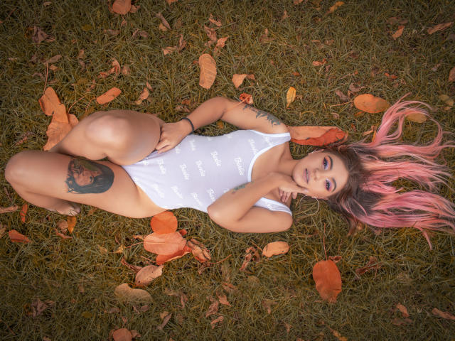 ValerieMartens con i capelli rosa, in un costume intero bianco, sdraiata sull'erba, con le gambe sollevate, che mostra un tatuaggio sulla coscia, circondata da foglie arancioni, sorride alla fotocamera, con le mani vicino al viso..