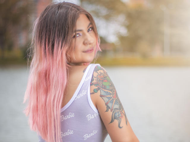 ValerieMartens, con i capelli rosa sfumati, una canottiera bianca, un braccio tatuato e un sorriso carino, guarda oltre la spalla, mostrando la schiena e i tatuaggi sul braccio superiore..