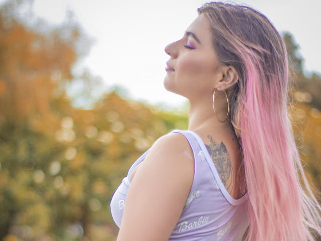 ValerieMartens, con i capelli rosa, orecchini a cerchio e ombretto viola, vista di profilo, che mostra un tatuaggio sulla parte superiore della schiena, indossa una canotta bianca, all'aperto, in una luce naturale soffusa..