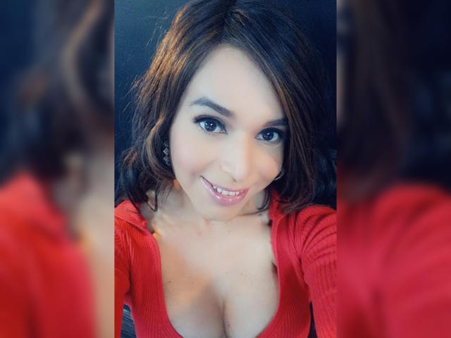 Jhovanna with medium brown wavy hair, red cardigan, and visible cleavage, smiling at her reflection in a mirror. Her tits are slightly exposed, and she has a cute face.