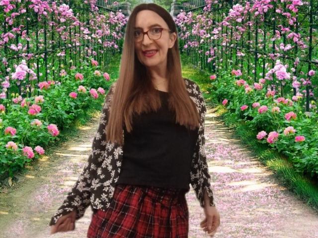 Emilianne a de longs cheveux bruns, des lunettes, un cardigan noir à motifs floraux, un haut noir et une jupe rouge à carreaux. Elle se tient debout sur un chemin rose bordé de fleurs, souriante..