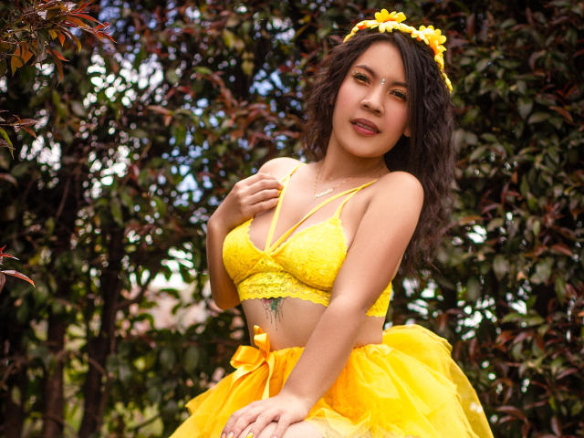 Helen Ross, con i capelli castano ricci, una fascia gialla con fiori, un reggiseno giallo in pizzo e una gonna gialla, tiene le mani sul petto, mostrando un piccolo tatuaggio verde sotto il seno..