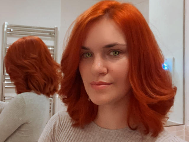LaraFontaineX with red hair, green eyes, and a nose ring, wearing a grey ribbed shirt, reflected in a mirror, showing her back. She's looking at the camera.