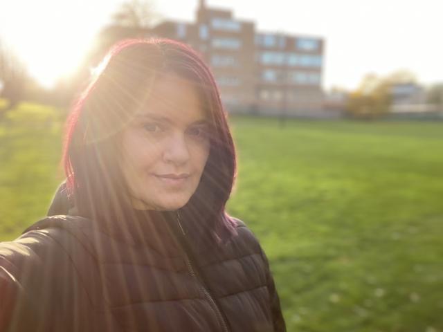 LaraFontaineX with pinkish-red hair, wearing a black puffy jacket, sunlight in face, green grass, and a blurred brick building in the background, smiling softly.