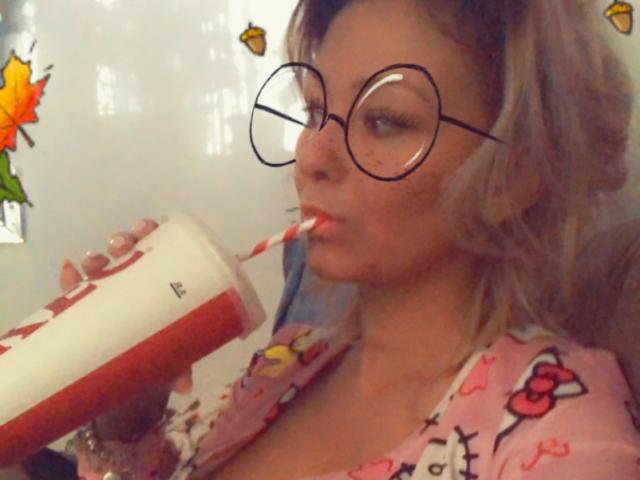 SpicyAlicia, com óculos redondos, bebendo de uma caneca vermelha e branca, veste uma camisa rosa com estampas de desenhos animados, tem cabelo loiro e está chupando um canudo listrado, parecendo adorável..