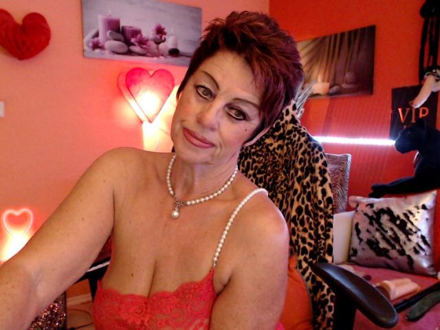 Bettina mit kurzen, roten Haaren, die ein rotes Spitzenoberteil und eine Perlenkette trägt, zeigt ihre Brüste und sitzt in einem Stuhl mit Leopardenmuster, wobei im Hintergrund Herz-Dekorationen zu sehen sind..