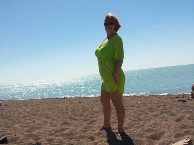 Anita, una fantasía con cabello rojo, gafas de sol y un vestido transparente verde neón, de pie en una playa arenosa, con un océano azul de fondo, usando sandalias rosas y mirando ligeramente hacia un lado..