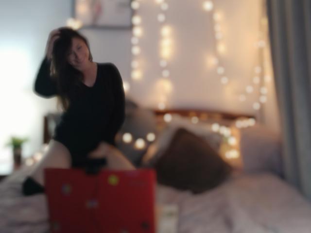 ValerySky with long brown hair, black long-sleeve top, and exposed thighs, kneeling on bed, hand in hair, blurred bokeh lights in background, red laptop in foreground.
