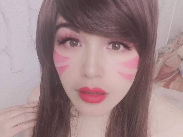 HikaruChan a des rayures de maquillage rose sur le visage, du rouge à lèvres rouge, et des cheveux bruns avec une frange, et elle regarde l'appareil photo avec ses yeux bruns. Sa main est près de son épaule..