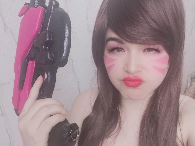 HikaruChan mit rosiger Gesichtsbemalung, rotem Lippenstift und langen braunen Haaren hält eine pinke und schwarze Wasserpistole. Ihre Brüste sind teilweise sichtbar, und ihr Ausdruck ist ernst..
