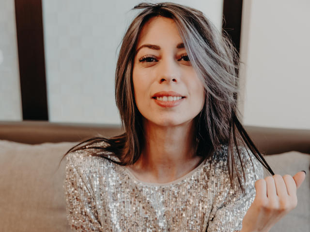 PoxyVibe with dirty blonde hair, sparkling silver sequin top, smiling, holding a strand of hair, soft light, cute, attractive, pretty, brown eyes, subtle makeup, casual, confident.