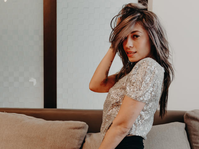 PoxyVibe with wavy brown hair, wearing a sparkly silver top, sits on a couch, hand in hair, looking over shoulder, smiling, showing a cute, sexy face.
