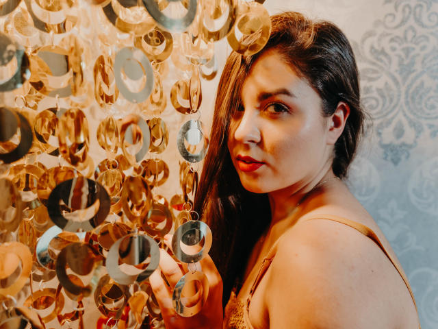 BelleInconnu with long brown hair, looking over her shoulder, holding a golden, circular, reflective, hanging object. She's wearing a yellow lace top, and her expression is slightly curious.