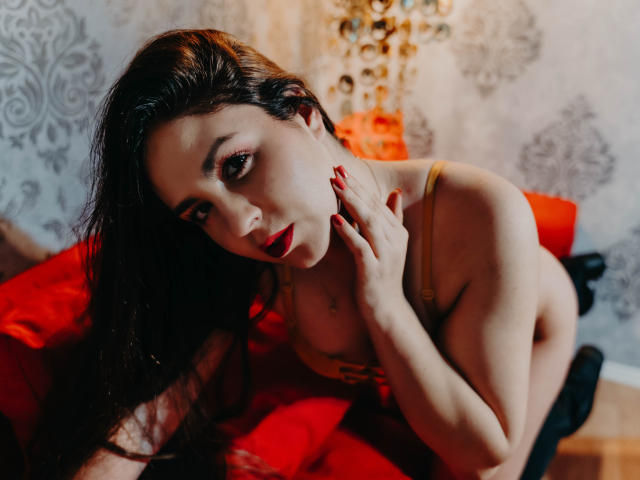 BelleInconnu with long dark hair, red lipstick, and yellow lingerie, touching her neck, leaning on a red couch, showing her tits, with red nails and a seductive gaze.
