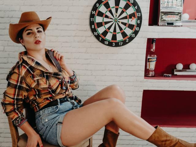BelleInconnu, with tan skin and dark eyebrows, wears a cowboy hat, plaid shirt, denim shorts, and brown boots, sitting with legs crossed, showing her bare thighs and small ass.