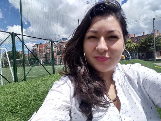 SakotoDika, com cabelos pretos ondulados, camisa branca com estampas, batom rosa e brincos de argola, sorrindo em um campo esportivo ao ar livre ensolarado. Ela tem seios de tamanho médio e um rosto bonito..