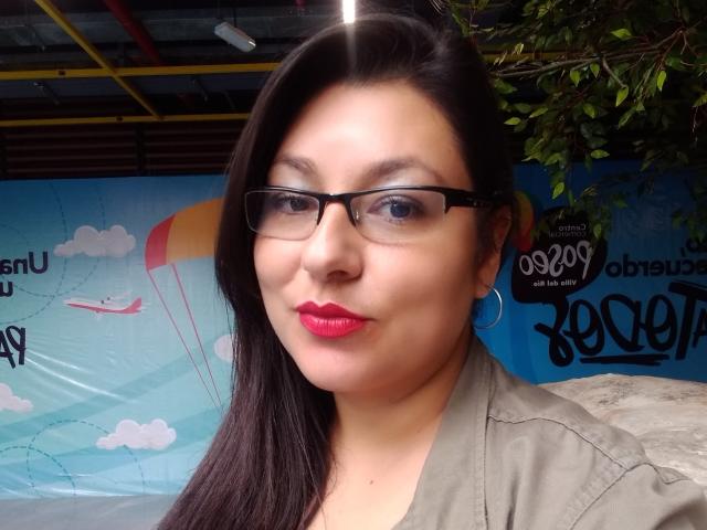 SakotoDika with red lipstick, black glasses, and long dark hair, wearing a green jacket, has a cute smile and is looking at the camera, showing her tits slightly.