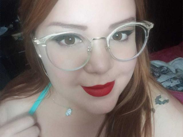 Fabiana, con rossetto rosso, occhiali e una tracolla color turchese, si tiene la spalla, mostrando un piccolo tatuaggio sulla spalla destra e una collana con un ciondolo..