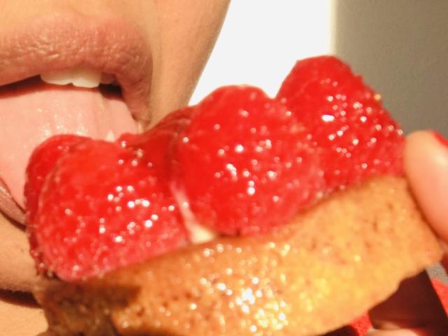 La lingua di PerleClara è fuori, mentre lecca una ciambella dorata con tre lampone rossi lucidi sopra. Le sue labbra rosa sono leggermente aperte..
