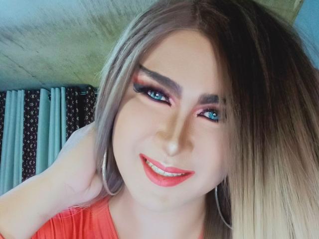 BornStarTS has blue eyes, long blonde hair, red lipstick, and is smiling at the camera, wearing a red shirt, with a hand in her hair.