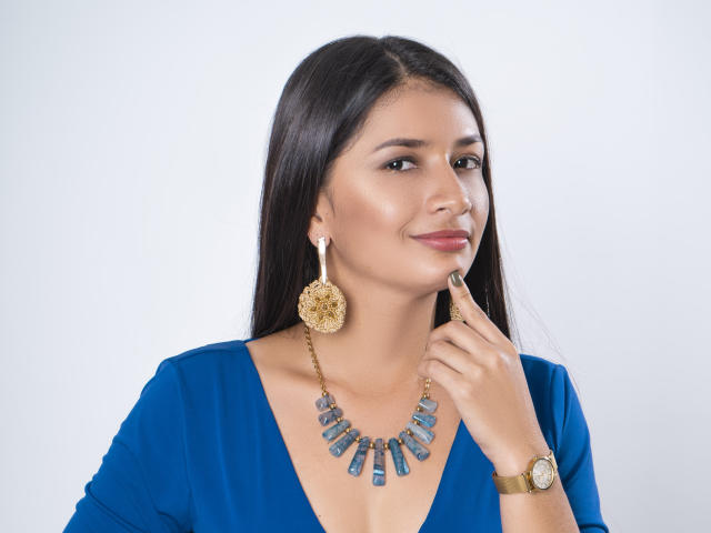 LeiaWhite tiene el cabello largo y oscuro, lleva un vestido azul, un collar dorado con piedras azules y grandes aretes dorados, toca su barbilla con un dedo y sonríe ligeramente..