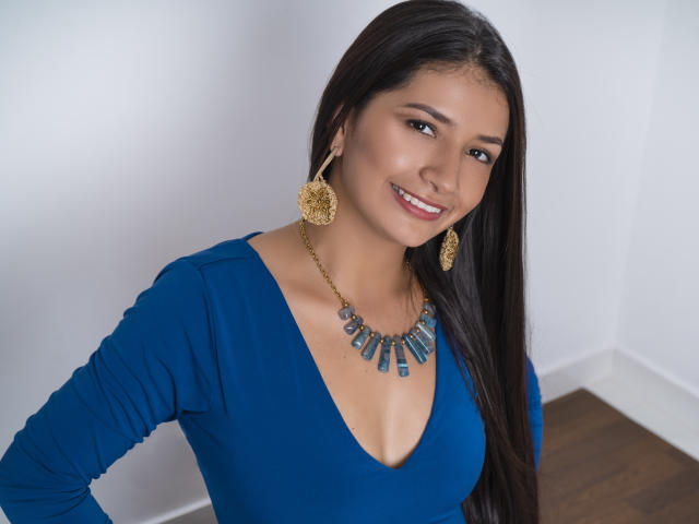 LeiaWhite, avec de longs cheveux noirs, une robe bleue, des boucles d'oreilles dorées et un collier de pierres bleues, souriant à la caméra, montrant sa poitrine, debout sur un plancher en bois, avec un mur blanc en arrière-plan..