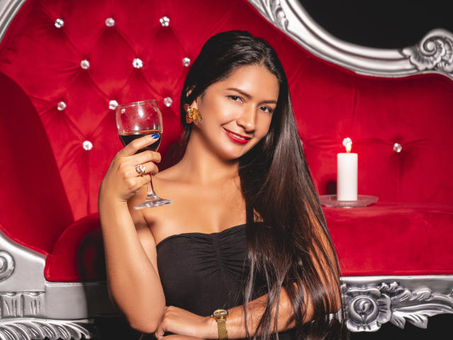 LeiaWhite met lang zwart haar, lachend, met een glas rode wijn in haar hand, gekleed in een zwarte jurk zonder bandjes, met gouden oorbellen en een ring, zittend op een rode fluwelen stoel..