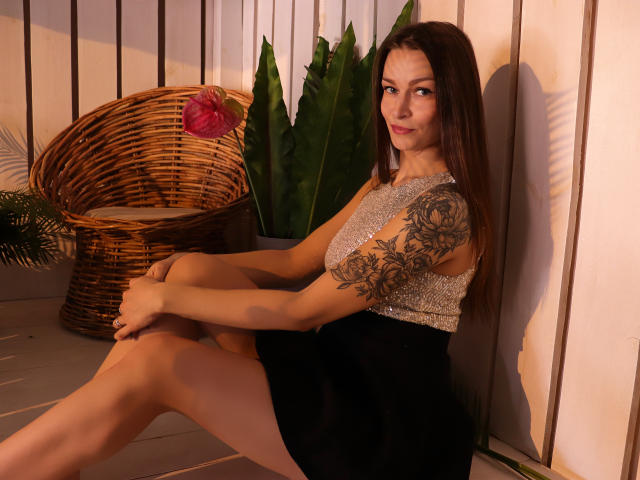 AlexiaBlack, with long brown hair, sitting on a wooden floor, wearing a sparkly sleeveless top and black skirt, showing a tattooed arm, and smiling at the camera.