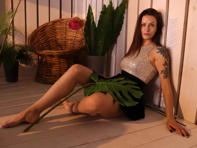 AlexiaBlack, con lunghi capelli castani, indossa una top scintillante senza maniche e una gonna verde a forma di foglia, seduta su un pavimento di legno, tiene in mano una grande foglia, mostra un tatuaggio sul braccio destro, piedi nudi..