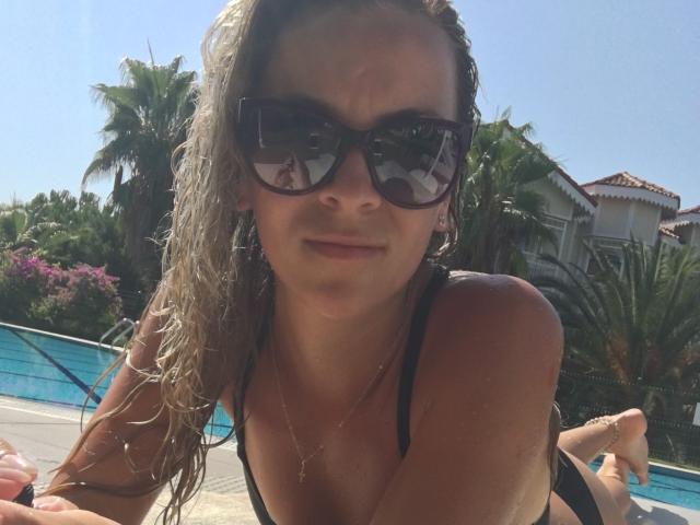 SophieDeee with wet blonde hair, black sunglasses, and black bikini top, lying by the pool, looking at camera, subtle smile, tan skin, visible tan lines, necklace, sunny day.