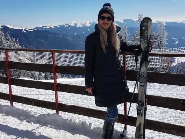 SophieDeee stands in snow, wearing a black puffy coat, blue jeans, black boots, and a beanie, holding skis with snow-covered mountains in the background.