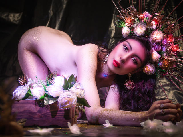 IsaTimeSweet is naked, bent over with sparkling skin, wearing a flower crown and star earring. She has red lipstick, dark eyes, and white roses in a vase below.