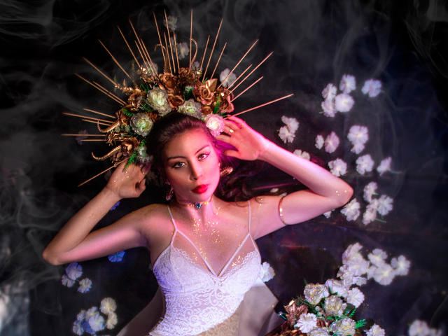 IsaTimeSweet trägt ein weißes Spitzenkleid, roten Lippenstift und eine goldene Krone mit Zacken, liegt in einer rauchigen, blumenreichen Szene und hält Blumen in der Nähe ihrer Brüste, wobei sie verführerisch wirkt..