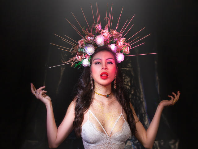 IsaTimeSweet, con cabello oscuro y ondulado, vestida con una blusa blanca de encaje y una corona floral rosa con espinas rojas, lápiz labial rojo, collar dorado, y con las manos levantadas, luciendo seductora..