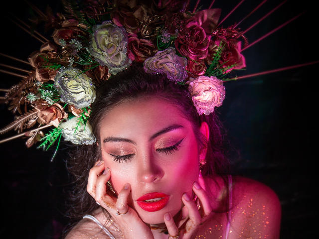 IsaTimeSweet avec une coiffe ornée de roses, du rouge à lèvres rouge, et des paillettes sur la peau, les mains touchant le visage, un fond sombre, un éclairage rose et rouge, des yeux doux, et un maquillage détaillé..