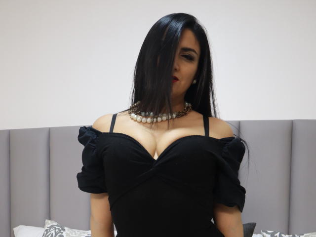 JuliaRobinson with black hair, wearing a black off-shoulder dress, showing big tits, necklace, and red lipstick, standing in front of a gray headboard.