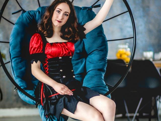 ClaptonFow mit welligem, braunem Haar, trägt ein rotes und schwarzes Korsettkleid und sitzt in einem türkisfarbenen, runden Stuhl. Die Brüste sind klein, die Brustwarzen sind wohlgeformt, die Vulva ist nicht sichtbar..