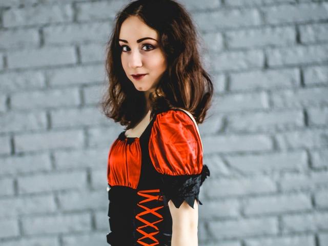 ClaptonFow, avec ses cheveux noirs et ondulés, portant une robe corset orange et noire, regardant par-dessus son épaule avec un léger sourire, debout devant un mur de briques grises..
