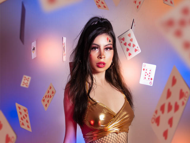 IsaTimeSweet, con su larga cabellera negra, top metálico dorado, medias de rejilla y maquillaje rojo, rodeada de cartas flotantes, tiene un busto grande y una mirada sexy y segura..