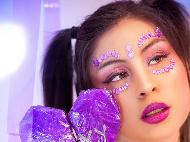 Das Gesicht von IsaTimeSweet, mit lila Glitzerherzmuster auf Stirn und Augenlidern, pinkem und lila Eyeliner, pinkem Lippenstift, lila, zerknittertem Plastik im Vordergrund, braunen Zöpfen, Blick zur Seite..