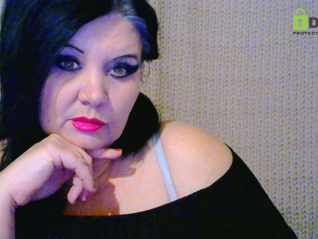 KamilaDream with black hair, green eyes, and red lipstick, wearing an off-shoulder black top, showing her white bra strap, resting her chin on her hand.