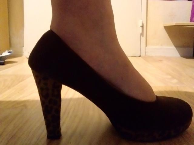 Le pied de LoveClo, chaussé d'une escarpin noir à imprimé léopard sur le talon, se tient sur un plancher en bois, montrant un profil latéral avec un léger angle ascendant..