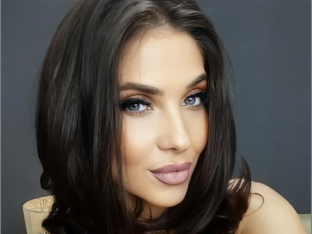 LovelyKinsley has dark, shiny hair, blue eyes, and pink lipstick, with a subtle smile. Her eyebrows are thick, and she has a smooth, fair complexion.