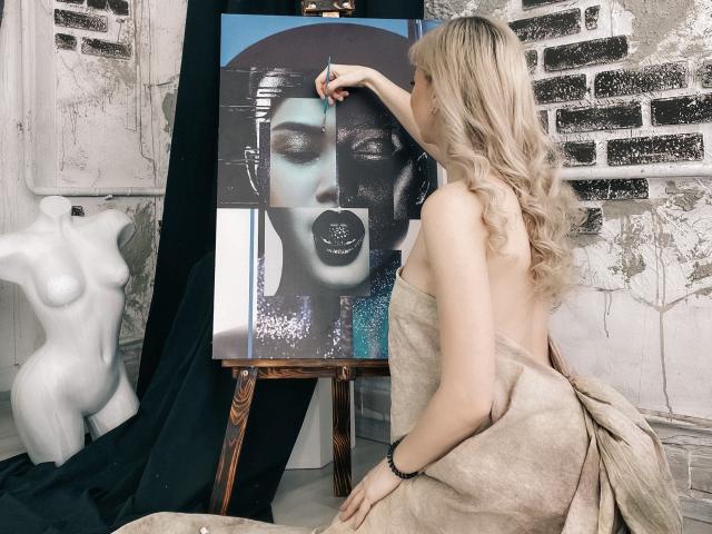 ParadiseRoxanne with blonde hair, back to camera, painting black-and-white portrait of woman with dark lipstick, using blue brush, wearing strapless beige dress, white nude bust on left.